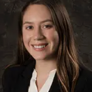 A headshot of a person with long brown hair, wearing a black blazer and white shirt, smiling in front of a dark textured background.