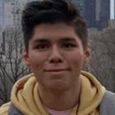 Headshot of a person wearing a yellow hoodie and backpack, with a cityscape in the background. Trees and buildings are visible.