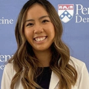 Headshot of a smiling person with long hair, wearing a white coat, standing in front of a backdrop with a Penn Medicine logo.