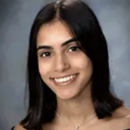 A headshot of a person with shoulder-length dark hair, smiling, wearing an off-the-shoulder top and a necklace, against a neutral background.