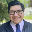 A person wearing glasses and a tie, smiling outdoors near greenery.