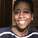 Headshot of a person smiling, wearing a dark shirt with white stripes, featuring makeup with defined eyeliner. The background is dimly lit.