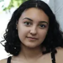 Headshot of a person with curly hair, standing outdoors against a wall with greenery in the background.