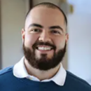 Headshot of a smiling person with a beard, wearing a blue sweater over a white collared shirt, in an indoor setting.