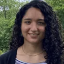 A headshot of a person with curly dark hair, wearing a striped top and a black cardigan, set against a backdrop of green foliage and a cloudy sky.