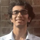 A headshot of a person wearing glasses and a light-colored shirt, smiling, with a brick wall in the background.