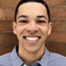 Headshot of a person smiling in front of a brick wall, wearing a collared shirt.