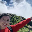 A person wearing a red sweatshirt and carrying a backpack is smiling and holding out an arm against a backdrop of green hills and a cloudy sky.