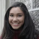 A person smiling in front of a textured stone wall, wearing a dark top.