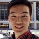 A person taking a selfie outdoors, wearing a checkered shirt. A building with large windows is in the background.