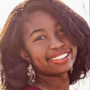 Headshot of a person with shoulder-length dark hair, smiling, wearing a red top and dangling earrings. τους.