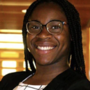 A headshot of a person smiling, wearing glasses and braided hair, dressed in a dark jacket over a patterned shirt, with a blurred indoor background.