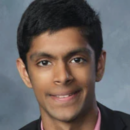 A person in a suit and pink shirt, smiling in a professional headshot against a gray background.