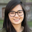 A headshot of a person with long dark hair and glasses, smiling, set against an outdoor background.