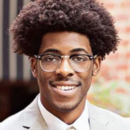 A headshot of a person with an afro hairstyle and glasses, wearing a light gray suit and a striped tie, smiling against a blurred outdoor background.