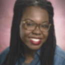 A headshot of a person wearing glasses, with long braided hair and a denim shirt, smiling against a pink background.