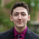 Headshot of a person wearing a dark suit, magenta shirt, and striped tie, set against a blurred outdoor background.