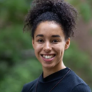 Headshot of a person with curly hair, smiling outdoors, wearing a black top. Background is blurred greenery.