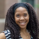 Headshot of a person with long curly hair, wearing a black and white striped top, smiling outdoors with a blurred background.