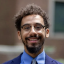 A headshot of a person with glasses, a beard, and curly hair, wearing a blue shirt, tie, and dark blazer, smiling in an outdoor setting.