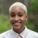 Headshot of a smiling person with short blonde hair, wearing earrings and a light-colored shirt, set against a blurred outdoor background.