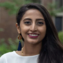 A person with long, dark hair smiles while wearing a white top and large earrings, with a blurred outdoor setting in the background.