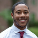 Headshot of a person smiling, wearing a light dress shirt and a red tie, with a blurred outdoor background.