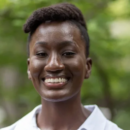 A headshot of a person smiling, wearing a white shirt, with a blurred greenery background.