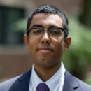 Headshot of a person wearing glasses, a dark suit, white shirt, and striped tie, against a blurred outdoor background.