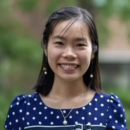 A person smiling in a headshot, wearing a blue polka dot top and pearl earrings, with a blurred outdoor background.