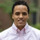 A headshot of a person wearing a light-colored, checkered button-up shirt, standing outdoors with a blurred natural background.