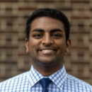 A headshot of a person smiling, wearing a blue tie and a blue and white checkered shirt, with a brick wall in the background.