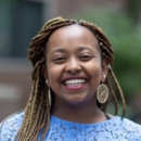 Headshot of a person smiling, wearing a blue lace top and large round earrings, with braided hair. The background is blurred.