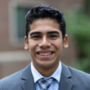 A headshot of a person smiling, wearing a gray suit and blue plaid tie, with a blurred outdoor background.