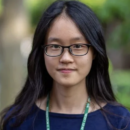 A person with long dark hair and glasses is wearing a navy shirt and a lanyard. The background is a blurred outdoor setting with greenery.