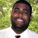 A headshot of a person smiling, wearing a white shirt and dark red tie, with a background of green foliage and tree branches.