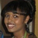 A headshot of a person smiling, with straight hair styled in a bun and bangs. The individual is wearing earrings and a green top.