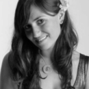 A black and white headshot of a person with long hair, wearing a flower in their hair and a necklace, smiling slightly.