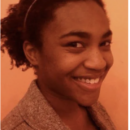 Headshot of a smiling person with curly hair, wearing a headband and a textured jacket, set against an orange background.