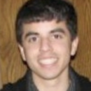 Headshot of a person with short dark hair, smiling, wearing a dark top, in front of a wooden background.