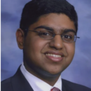 Headshot of a person wearing glasses, a suit, and a tie, smiling against a blue background.