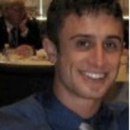 A headshot of a person smiling, wearing a blue shirt and tie, with tables and other people blurred in the background.