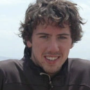 A headshot of a person with curly hair wearing a brown jacket, smiling outdoors with a slightly cloudy sky in the background.