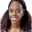 A headshot of a person smiling, wearing a necklace and a patterned top. The individual has braided hair.