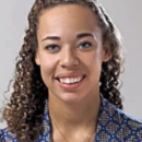 A headshot of a person smiling, with curly hair, wearing a blue patterned shirt, against a neutral background.