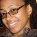 Headshot of a person smiling, wearing glasses and a zip-up jacket with a hood. Hair appears to be styled in braids or locs.