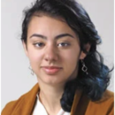 Headshot of a person with long dark hair, wearing a brown jacket and earrings, against a neutral background.