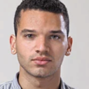 Headshot of a person with short, curly hair wearing a striped shirt. The person has earrings and a neutral expression.