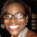 A close-up headshot of a person wearing glasses and smiling broadly. The background is dimly lit.