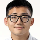 A headshot of a person wearing round glasses and a white shirt, with short, dark hair, smiling slightly against a plain background.